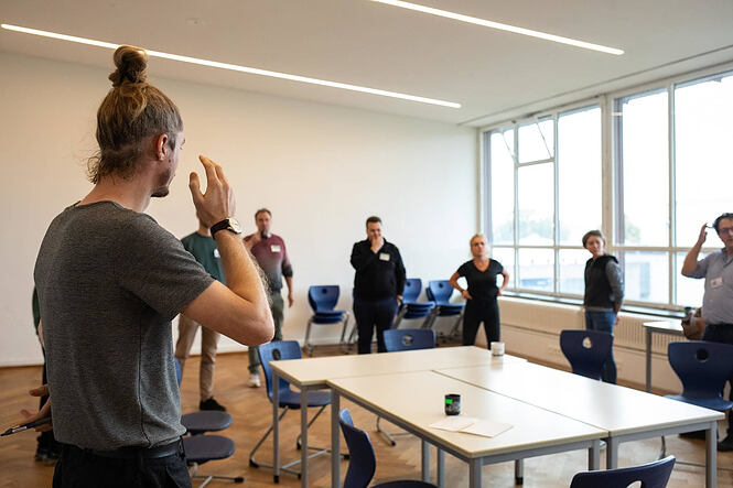 Das Bild zeigt eine Gruppe von Menschen in einem hellen Seminarraum mit großen Fenstern. Im Vordergrund steht eine Person mit zusammengebundenen Haaren und grauem T-Shirt, die offenbar spricht oder etwas erklärt und dabei eine Hand hebt. Mehrere Personen stehen im Hintergrund in lockerer Haltung, hören zu oder beobachten. Im Raum stehen einige Tische und blaue Stühle, teils im Kreis oder in kleinen Gruppen angeordnet. Das Licht ist natürlich, und die Atmosphäre wirkt konzentriert, aber entspannt – typisch für einen Workshop oder eine Gruppendiskussion.